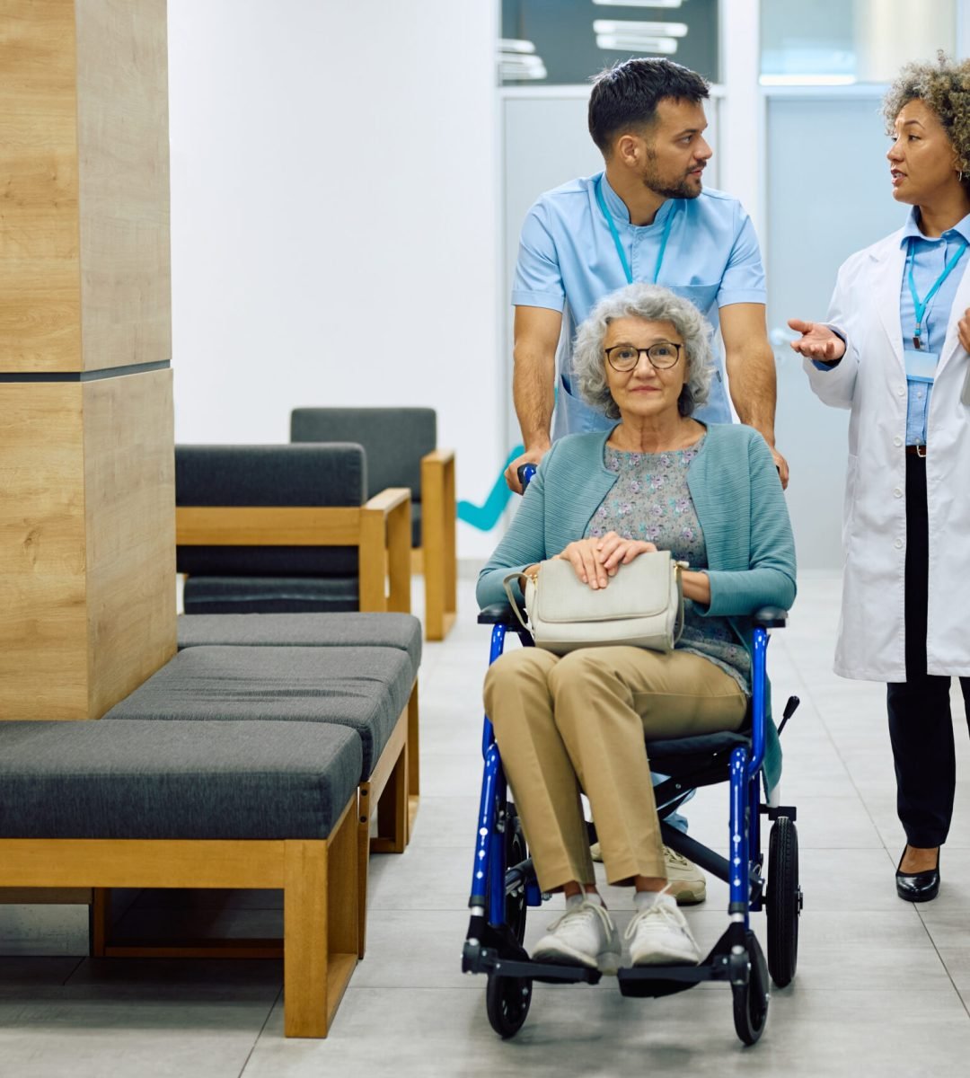 Female doctor communicating with medical technician who is pushing senior female patient in wheelchair at the clinic.