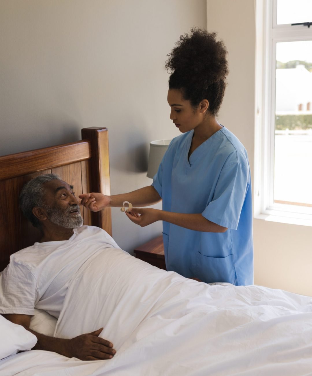 Front view of a young African American female doctor fitting a senior African American man with hearing aid in bedroom at home